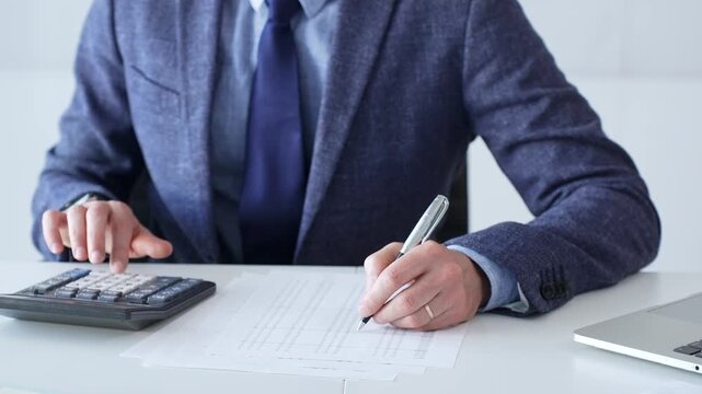 Businessman wearing suit and tie is using calculator and taking notes while working at desk in office, with laptop and stack of folders nearby, close up. Audit and taxes theme in business