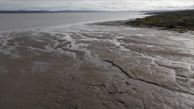 Cinematic nature landscape timelapse of tidal shoreline, muddy coastline, winding water channel, textured mudflat and tranquil background beneath overcast sky with distant coastal hills