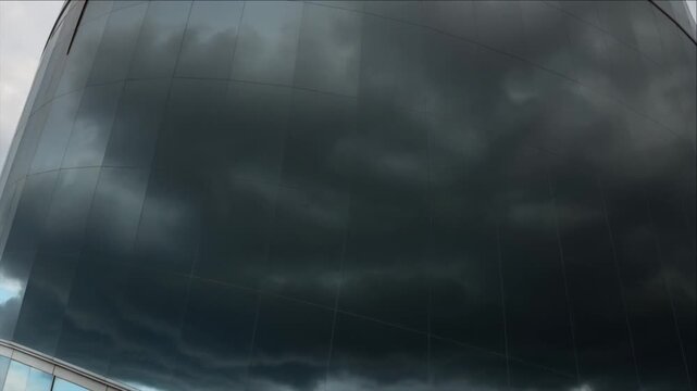 Cinematic urban weather timelapse background featuring dark storm cloud reflection across modern curved glass building facade, dramatic blue sky, moody architectural city atmosphere, searchable