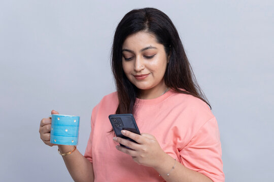 Portrait of an young Indian woman smiling while 
looking at her smartphone and holding a blue coffee mug isolated on gray studio background.
