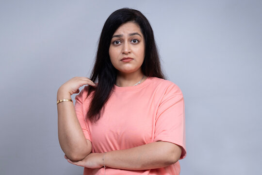 Young Indian woman in a pink casual shirt posing against a neutral gray studio background with folded arms and a pensive facial expression, representing concern, uncertainty and emotional.