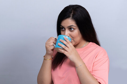 Young Indian woman drinking from a blue ceramic mug while looking to the side, wearing a pink casual shirt in a gray studio background.