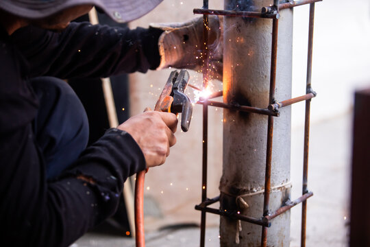 Selective focus to Male construction worker wearing sunglasses and bucket hat welding a steel structure at a job site. Glowing bright sparks fly off the electric metal arc welding process during outdo