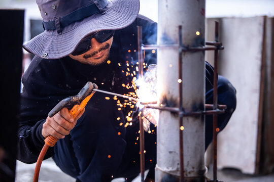 Selective focus to Male construction worker wearing sunglasses and bucket hat welding a steel structure at a job site. Glowing bright sparks fly off the electric metal arc welding process during outdo