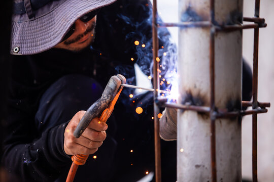 Selective focus to Male construction worker wearing sunglasses and bucket hat welding a steel structure at a job site. Glowing bright sparks fly off the electric metal arc welding process during outdo