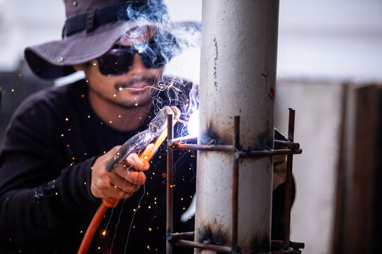 Selective focus to Male construction worker wearing sunglasses and bucket hat welding a steel structure at a job site. Glowing bright sparks fly off the electric metal arc welding process during outdo