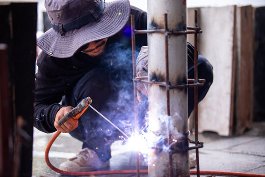 Selective focus to Male construction worker wearing sunglasses and bucket hat welding a steel structure at a job site. Glowing bright sparks fly off the electric metal arc welding process during outdo