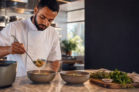 Male chef ladling hot soup into ceramic bowls beside fresh herbs in a sunlit restaurant kitchen