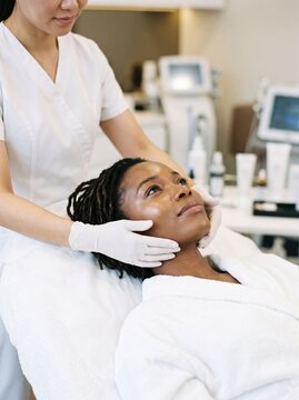 African American woman with locs lies relaxed as a gloved esthetician performs a facial treatment, a skincare and spa wellness concept