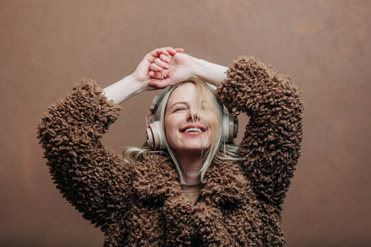 A stylish blonde woman wearing a cozy Teddy coat paired with modern headphones, standing against a rich brown background in shade Mocha Mousse