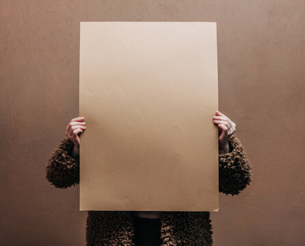 A stylish blonde woman wearing a teddy coat with paper,  standing against a rich brown background in shade Mocha Mousse