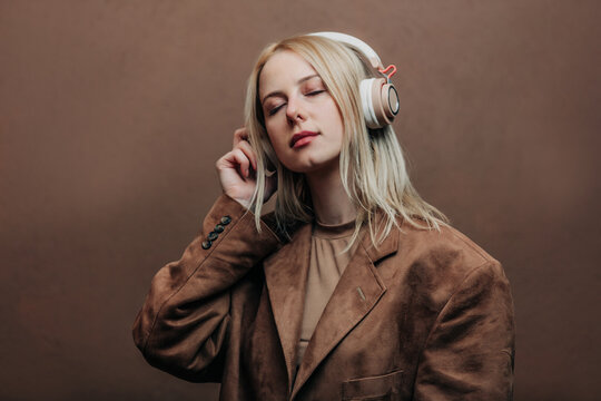 A stylish blonde woman wearing a brown jacket paired with modern headphones, standing against a rich brown background in shade Mocha Mousse