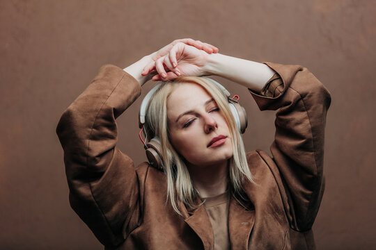 A stylish blonde woman wearing a brown jacket paired with modern headphones, standing against a rich brown background in shade Mocha Mousse