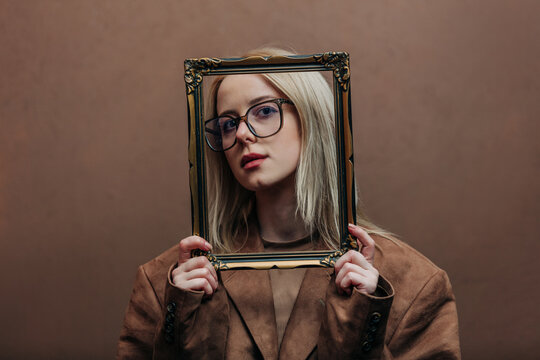 A stylish blonde woman wearing a brown jacket paired with vintage picture frame, standing against a rich brown background in shade Mocha Mousse