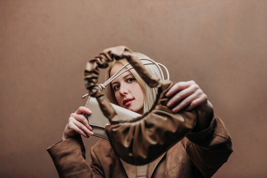 A stylish blonde woman wearing a brown jacket paired with vintage bags, standing against a rich brown background in shade Mocha Mousse