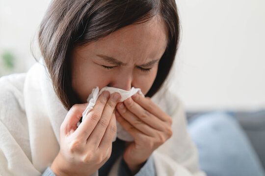 Sick Asian woman blowing her nose with tissue on sofa, wrapped in blanket, suffering from high fever, seasonal flu, influenza virus infection, and cold symptoms at home