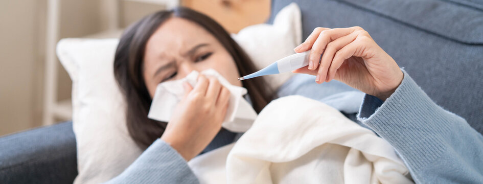 Sick person resting in bed checking temperature with thermometer, suffering from high fever, seasonal flu, influenza virus infection, and common cold symptoms at home