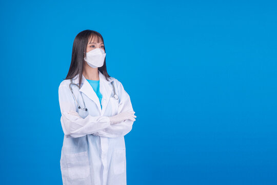 Confident young Asian female doctor wearing a white face mask, stethoscope, and lab coat standing with crossed arms on a blue background. Healthcare and medical protection concept.