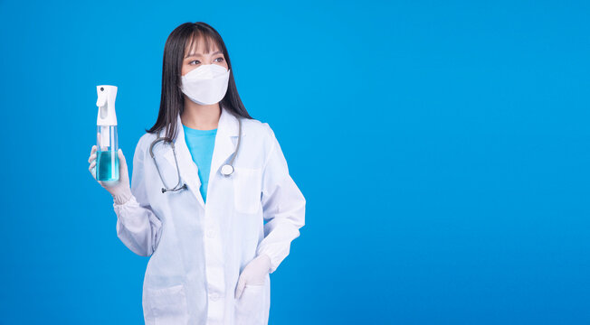 Confident young Asian female doctor wearing a face mask and lab coat holding an alcohol spray bottle on a blue background. Healthcare, hygiene, and virus protection concept. Perfect for medical ads.