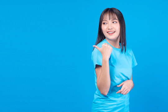 Happy smiling young Asian woman in blue t-shirt pointing thumb at empty copy space on blue background. Perfect for advertising, marketing, and presenting new promotion concepts.