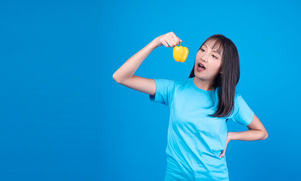 Playful young Asian woman in blue t-shirt holding a fresh yellow bell pepper and looking at it with open mouth on blue background. Healthy diet, vegan food, and organic nutrition concept.