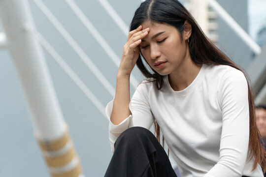 Sad young Asian woman touching her forehead feeling stressed and depressed while sitting alone outdoors in a modern city. Concept of mental health, headache, anxiety, and urban life pressure.