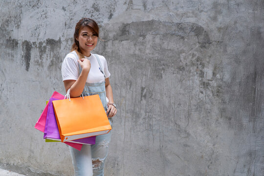 Happy young Asian woman in denim overalls holding colorful shopping bags and smiling against a grunge concrete wall background. Casual street fashion and retail shopping sale concept.