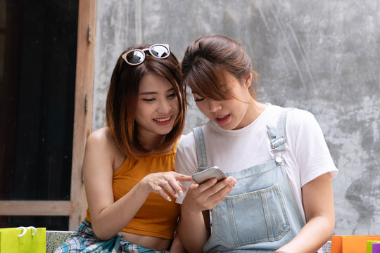 Two happy young Asian women friends looking at smartphone together and smiling while sitting outdoors with colorful shopping bags. Online shopping, social media, and retail lifestyle concept.