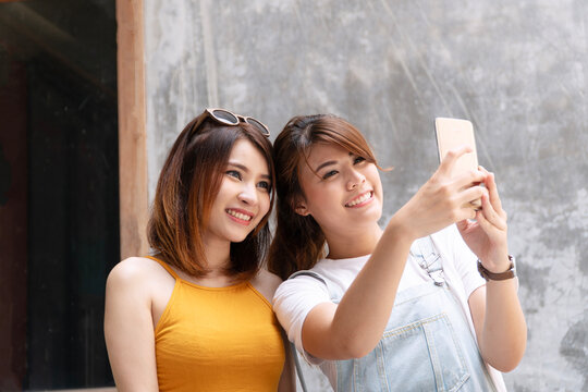Two happy young Asian women friends taking a selfie with a smartphone and smiling together against a grunge concrete wall. Social media, friendship, and mobile lifestyle concept.