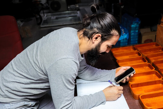 Side View Shows Bearded Man Leaning Over Table, Writing In Notebook While Holding Smartphone Near Rows Of Orange Storage Trays, Suggesting Planning, Inventory Review, Or Detailed Task