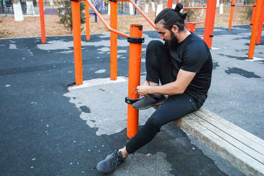Man In Black Athletic Clothing Sits On Wooden Bench At Outdoor Fitness Park, Tying Running Shoe Beside Bright Orange Exercise Bars And Worn Paved Ground, Showing Focused Preparation During Workout