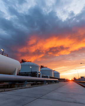 Sunset casts fiery hues over industrial tanks. Metal structures stand stark against the glowing sky. Concrete pathways lead toward distant horizons. Silent machinery reflects twilight's quiet glow