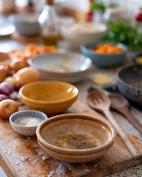 Warm kitchen bathed in golden afternoon light. Wooden bowls hold spices, herbs, and oil. Fresh onions, apples, and carrots add color. Cutting board shows flour dust and gentle use