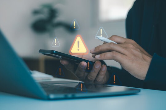 Businessman using smartphone and laptop while warning icons signal cybersecurity risk, digital threats, data security, online safety, and technology vulnerability in a modern workplace.