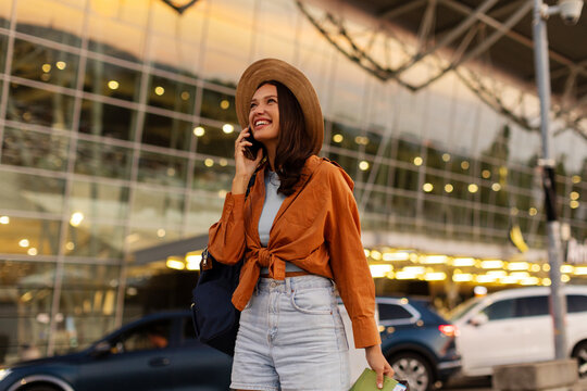 Happy woman traveling with backpack and suitcase, talking on cellphone while walking outside contemporary airport in the evening
