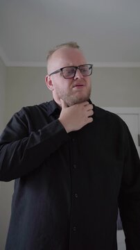 Adult man in glasses presses his throat and winces as swallowing causes discomfort against a plain indoor background.