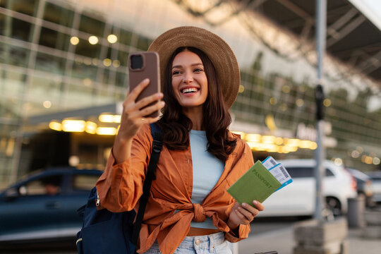 Happy woman holding passport with boarding passes while taking selfie or video calling with her smartphone outside airport terminal