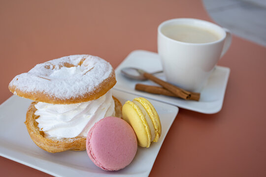 Close-Up Meringue Dessert with Pink and Yellow Macarons and Cappuccino. Close-up three-quarter view of an indulgent cream-filled meringue dessert on a white square plate, accompanied by pink and yello