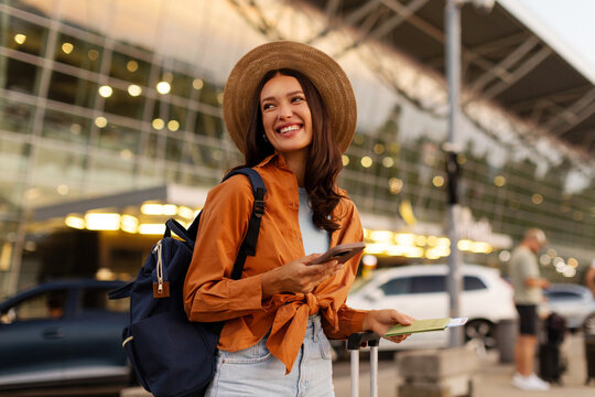 Excited lady ready for vacation, holding passport and flight ticket while checking her smartphone outside contemporary airport, looking aside and smiling