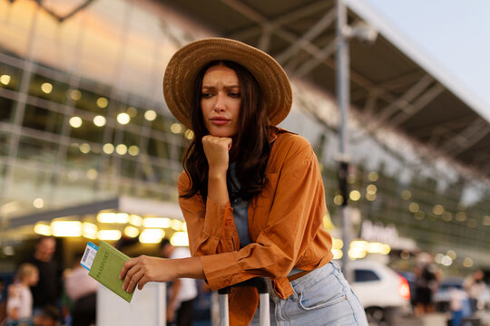 Worried woman leaning on her suitcase and holding passport standing outside airport, missed her flight and thinking