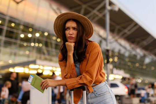 Sad woman holding passport and standing with her suitcase outside modern airport after her flight was canceled
