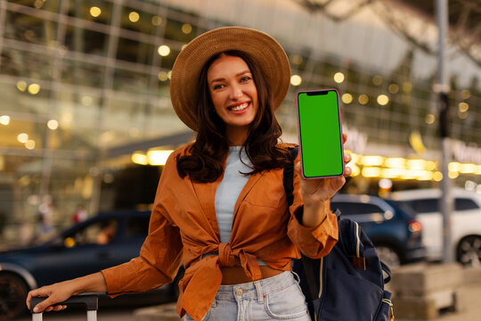 Smiling woman traveler showing blank smartphone screen while standing with suitcase and backpack outside modern airport terminal, mockup