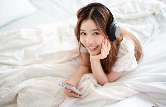 Mindfulness young adult asian woman lying in bed, wearing headphones listening relax music in cozy comfort bedroom