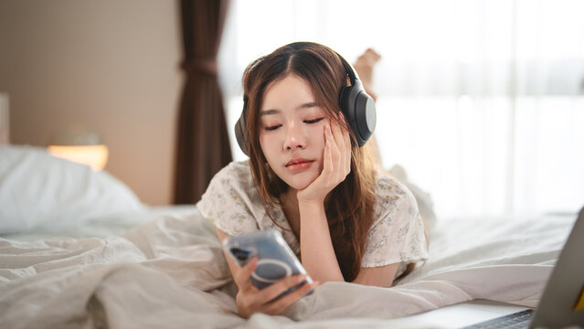 Mindfulness young adult asian woman lying in bed, wearing headphones listening relax music in cozy comfort bedroom