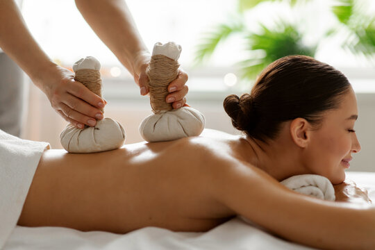 Happy female client receiving soothing herbal compress treatment on her back, experiencing muscle relief, stress reduction and complete body rejuvenation, side view