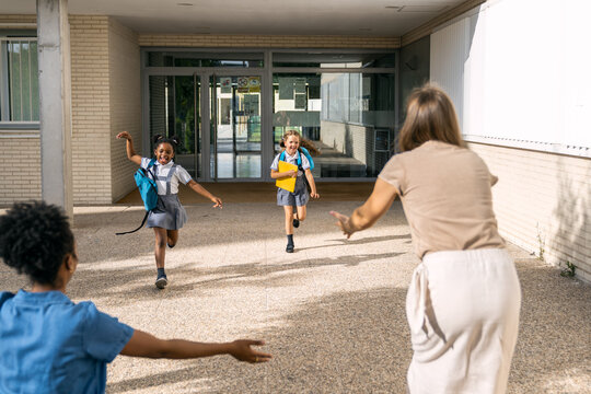 Happy multiracial girls running, embracing mothers at school entrance on their first day back, celebrating reunion and education