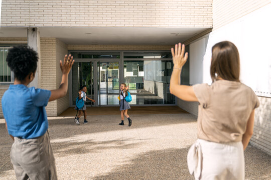 Multiethnic mothers waving goodbye to diverse primary students on their first day back to school, fostering friendship and community