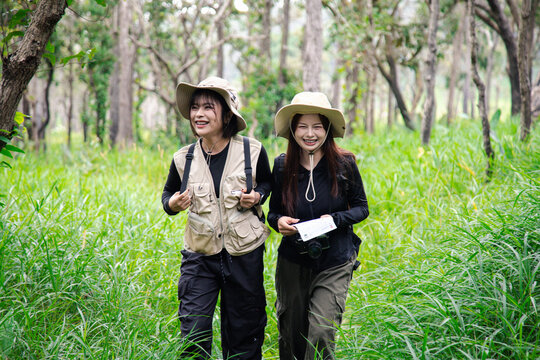 Two happy young Asian female friends hiking together in green forest, carrying backpacks, enjoying eco travel and nature.
