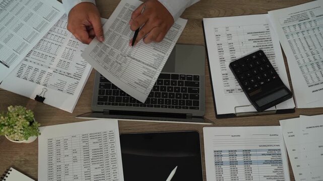 Accountant checking financial reports with pen beside laptop, calculator, tablet for accounting, tax preparation, audit, bookkeeping, financial planning.