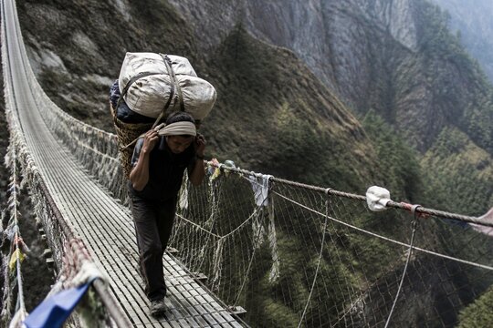 Highland porter carrying a heavy load across a suspended rope bridge above a mountain gorge. Documentary travel scene showing endurance, labor, transport and remote community life.